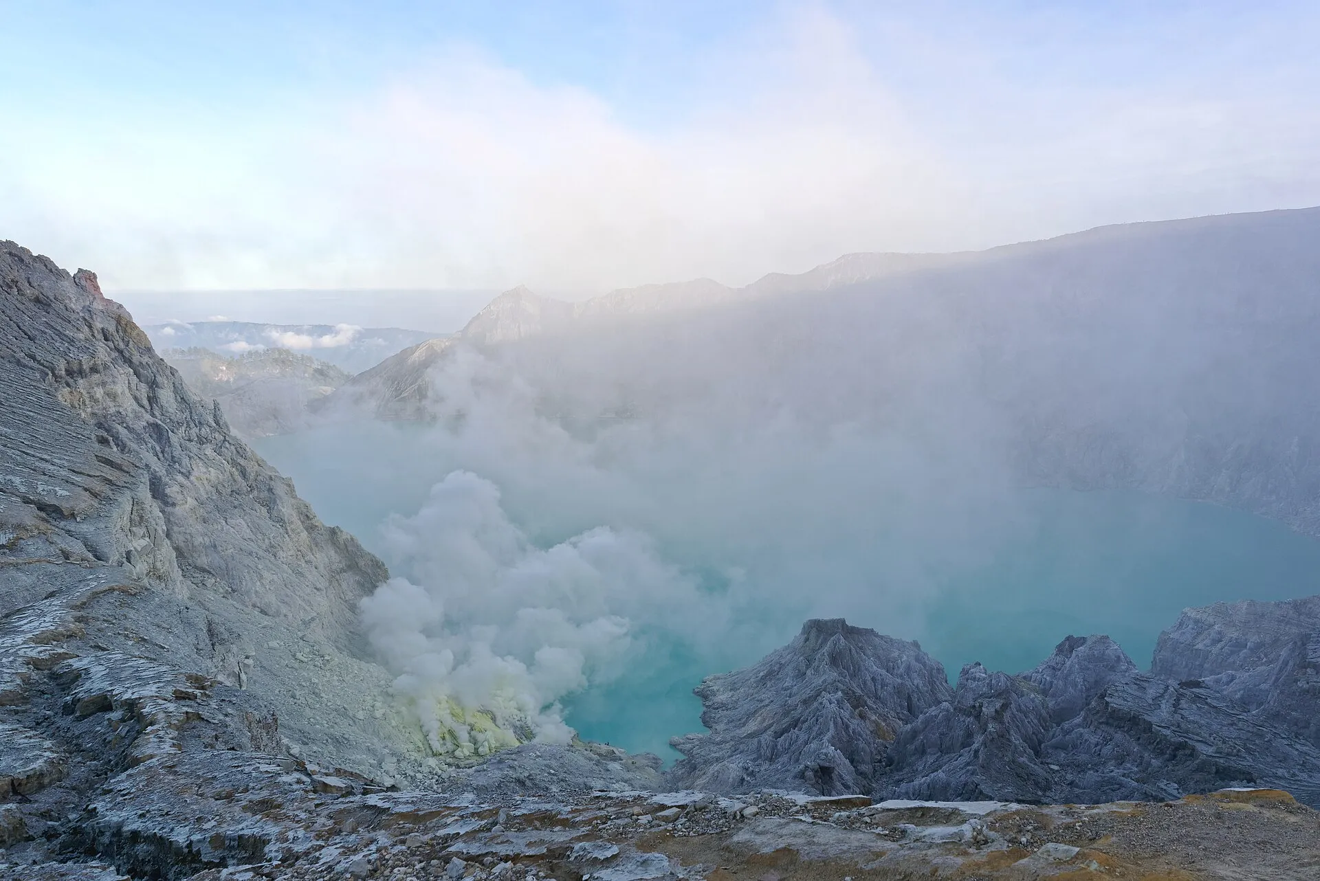 Danau kawah biru kehijauan di Kawah Ijen dengan dinding kawah vulkanik di sekelilingnya.