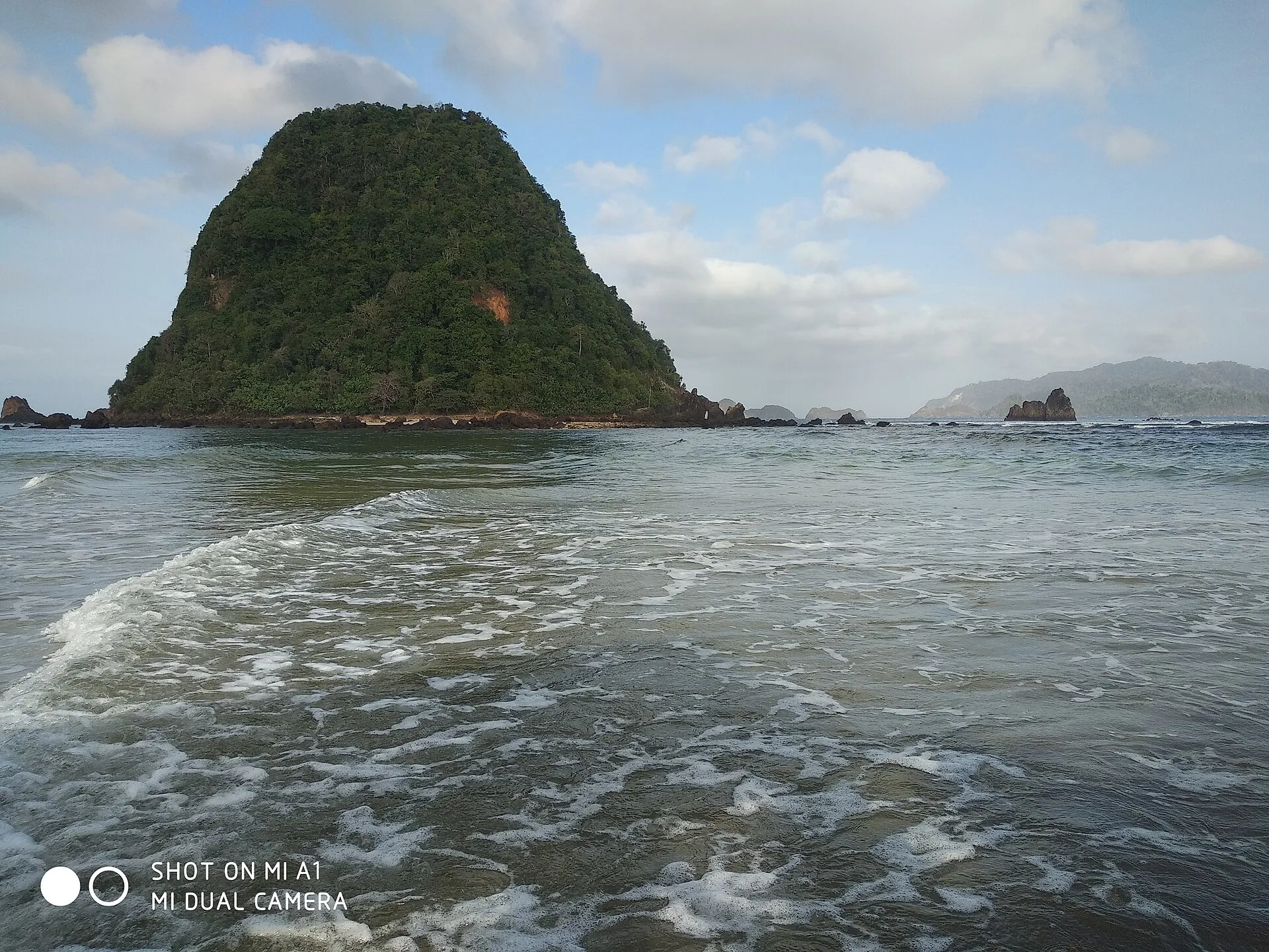 Pantai Pulau Merah dengan pasir gelap, laut biru, dan bukit kecil di kejauhan.